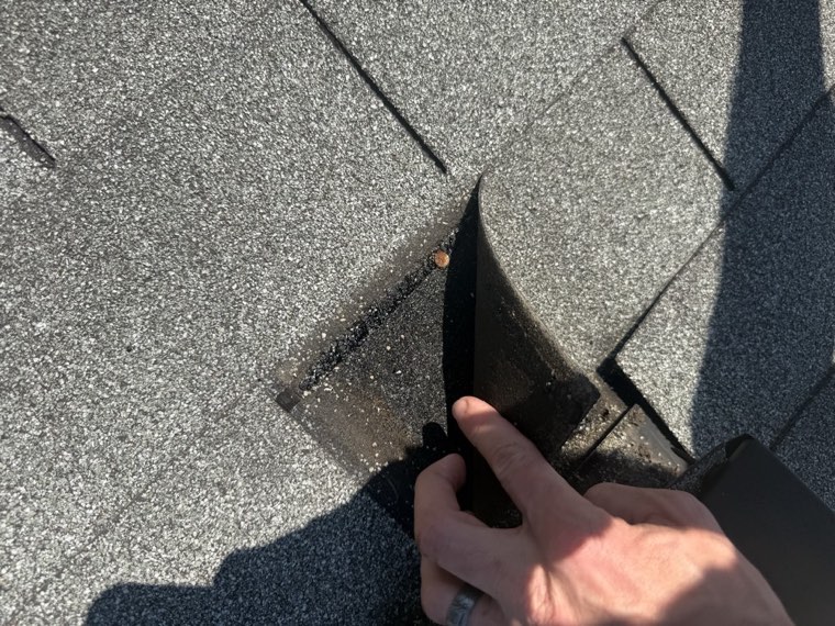 A lifted asphalt shingle pulled back to reveal an exposed nail and the gap where water gets in